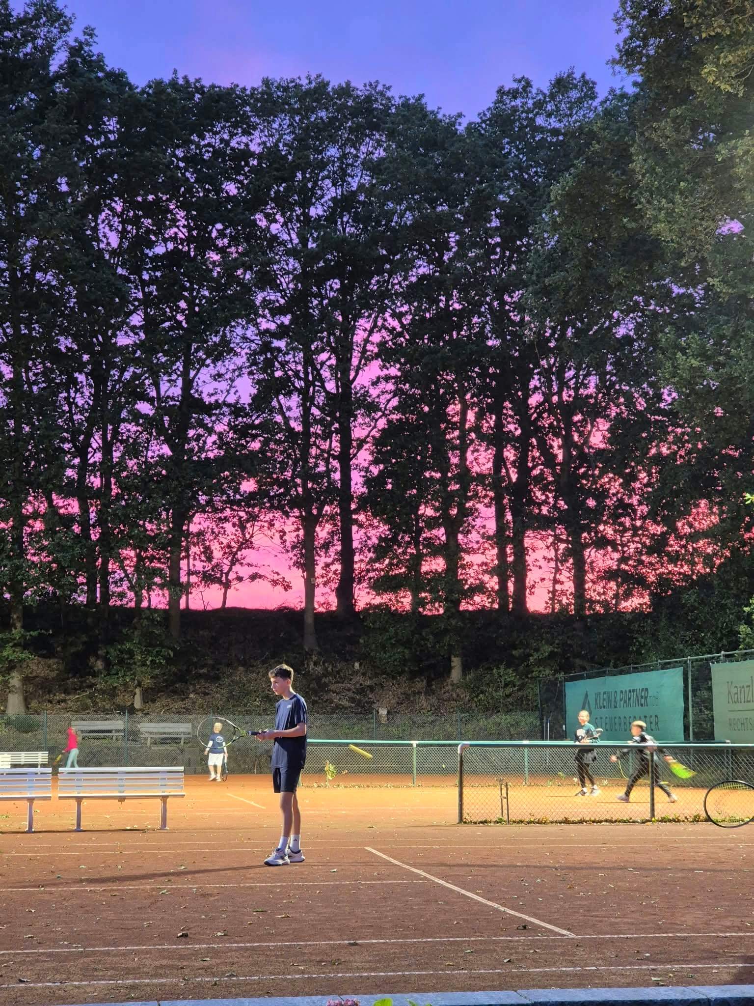 Jugendlicher Tennisspieler beim Flutlichtabend des Tennis-Club Oyten auf dem Sandplatz vor einem leuchtend pink-violetten Abendhimmel; im Hintergrund spielen weitere Jugendliche Tennis.